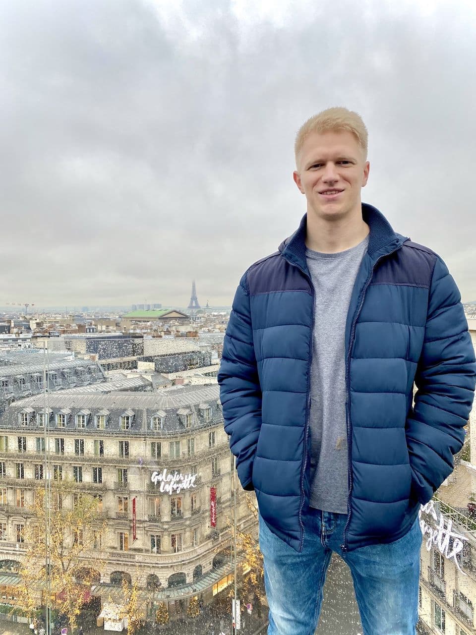 Luke on a rooftop in Paris, with the city's rooftops and the Eiffel Tower behind him on a gray day